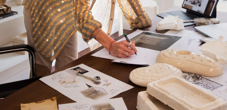 Persona escribiendo sobre bocetos en un escritorio con prototipos, muestras de biomateriales y documentos de diseño industrial.